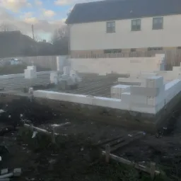 Wznoszone fundamenty domu z bloczków betonowych na przygotowanym terenie, widoczne drewniane szalunki i zbrojenie, w tle budynek mieszkalny i pochmurne niebo.