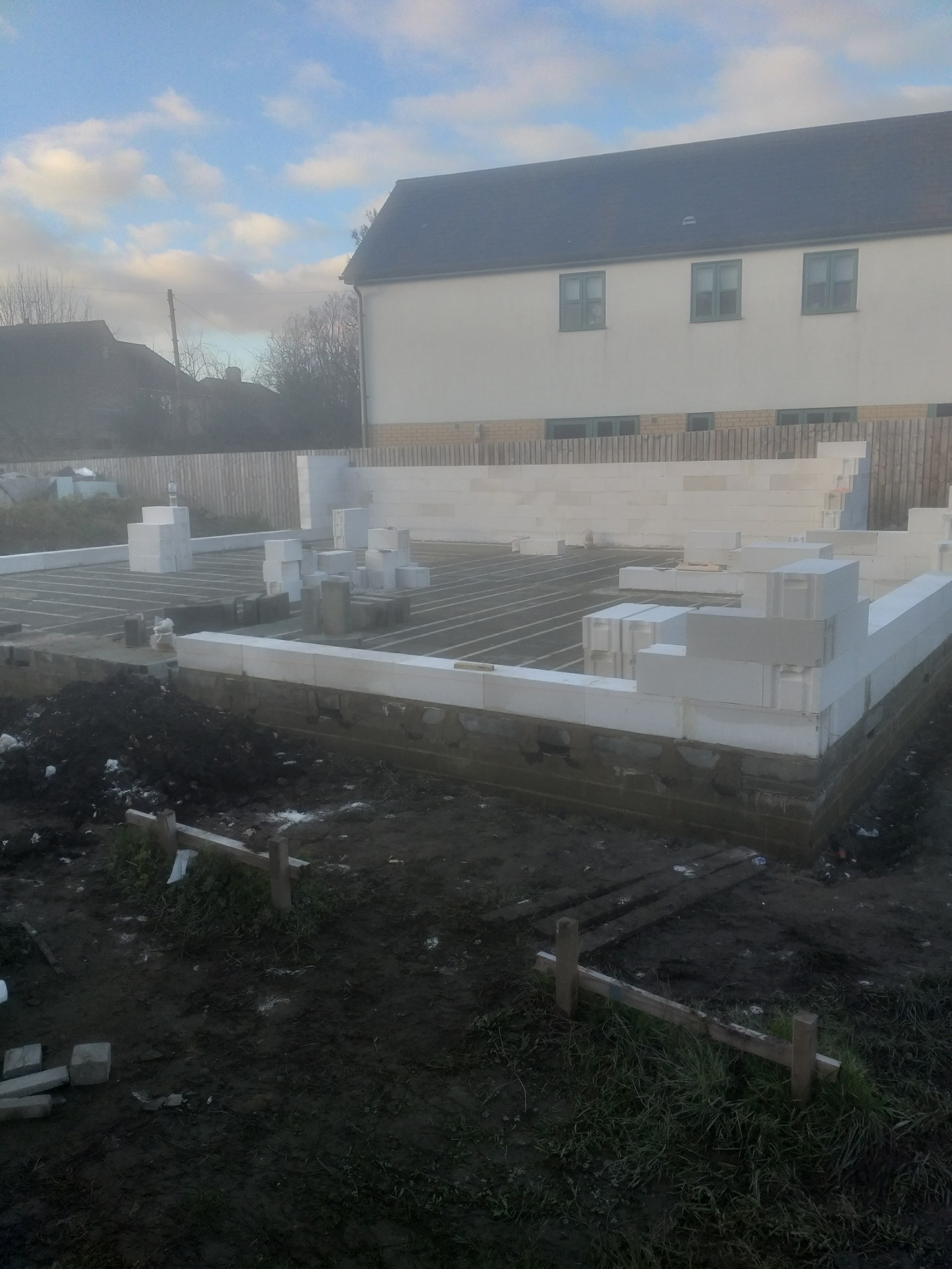 Wznoszone fundamenty domu z bloczków betonowych na przygotowanym terenie, widoczne drewniane szalunki i zbrojenie, w tle budynek mieszkalny i pochmurne niebo.