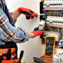 Elektryk w rękawicach ochronnych instaluje wyłącznik w skrzynce z bezpiecznikami, używając śrubokręta; obok leży multimetr cyfrowy.