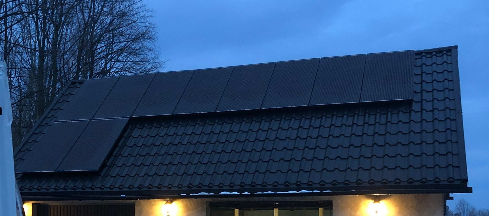 Panele fotowoltaiczne na dachu domu z ciemnej dachówki, widoczne o zmierzchu, z oświetleniem zewnętrznym.