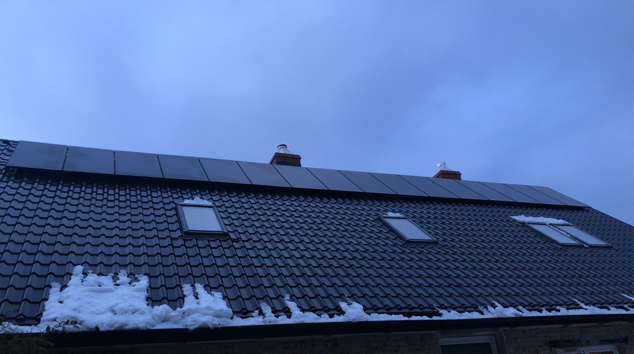 Panele słoneczne na ciemnym dachu z dachówkami, widoczne trzy okna dachowe i fragmenty śniegu u dołu dachu, pochmurne niebo.
