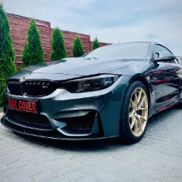 Grafitowe BMW M4 z przyciemnionymi szybami i lampami, złotymi felgami, emblematami Shadow Line i usuniętym chromem, zaparkowane na kostce brukowej przed drewnianym ogrodzeniem.