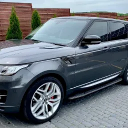 Szary Range Rover z przyciemnionymi szybami, zaparkowany na brukowanej nawierzchni, z widocznym samochodem firmowym w tle.