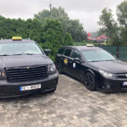 Dwa czarne samochody taxi zaparkowane na brukowanym podjeździe przed zielonymi tujami, z widoczną stacją Orlen w tle.