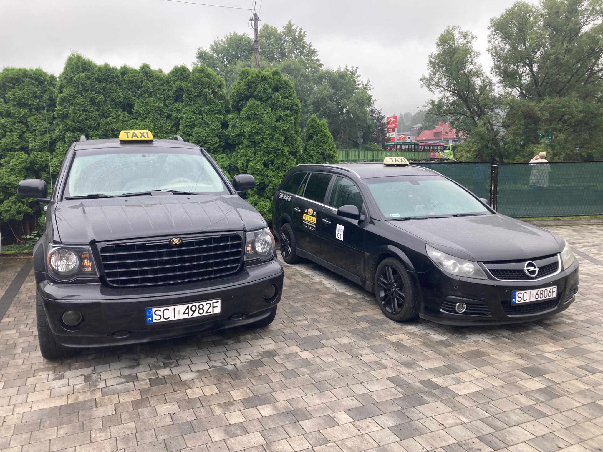 Dwa czarne samochody taxi zaparkowane na brukowanym podjeździe przed zielonymi tujami, z widoczną stacją Orlen w tle.