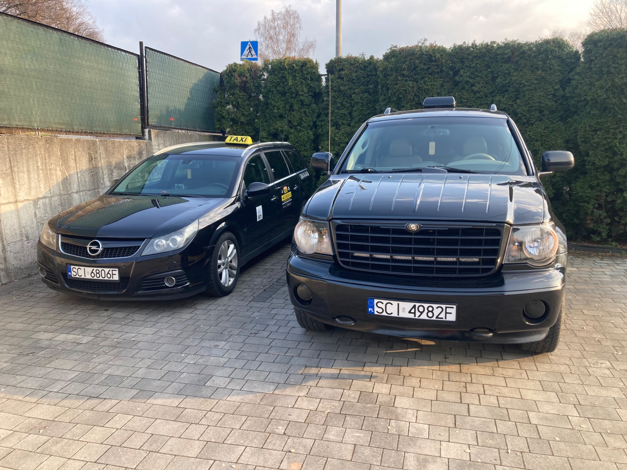 Dwa czarne samochody, w tym jeden z napisem TAXI na dachu, zaparkowane na brukowanym podjeździe przed zielonym żywopłotem i znakiem drogowym.