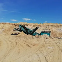 Praca przesiewacza do piasku w kopalni odkrywkowej, widoczne ślady opon na piasku i błękitne niebo.