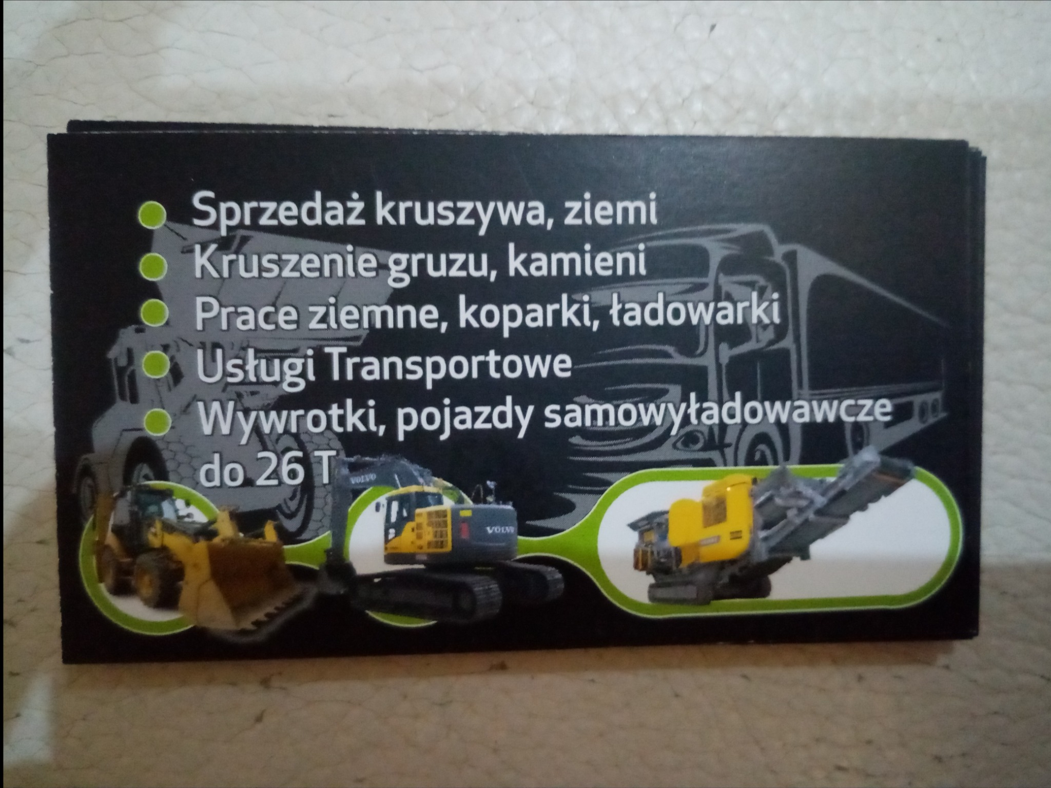 Wizytówka firmy oferującej sprzedaż kruszyw, kruszenie gruzu i kamieni, prace ziemne koparkami i ładowarkami oraz usługi transportowe wywrotkami do 26 ton. Na wizytówce grafika przedstawia...