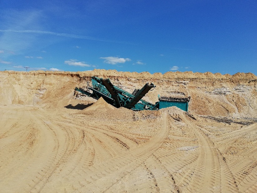 Praca przesiewacza do piasku w kopalni odkrywkowej, widoczne ślady opon na piasku i błękitne niebo.