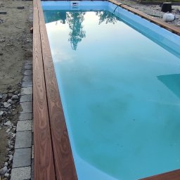 Elerk construction - Niebieski basen z drewnianym obramowaniem w trakcie budowy, widoczny pracownik i dom w tle.