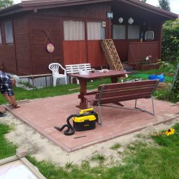 Elerk construction - Układanie czerwonej kostki brukowej na tarasie przed drewnianym domkiem letniskowym, widoczny pracownik w kraciastej koszuli, stół z ławkami i odkurzacz przemysłowy.