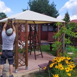 Elerk construction - Monterażysta przykręca wkrętarką element drewnianej pergoli z białym dachem na tle ogrodu z kwitnącymi rudbekiami i domkiem letniskowym.