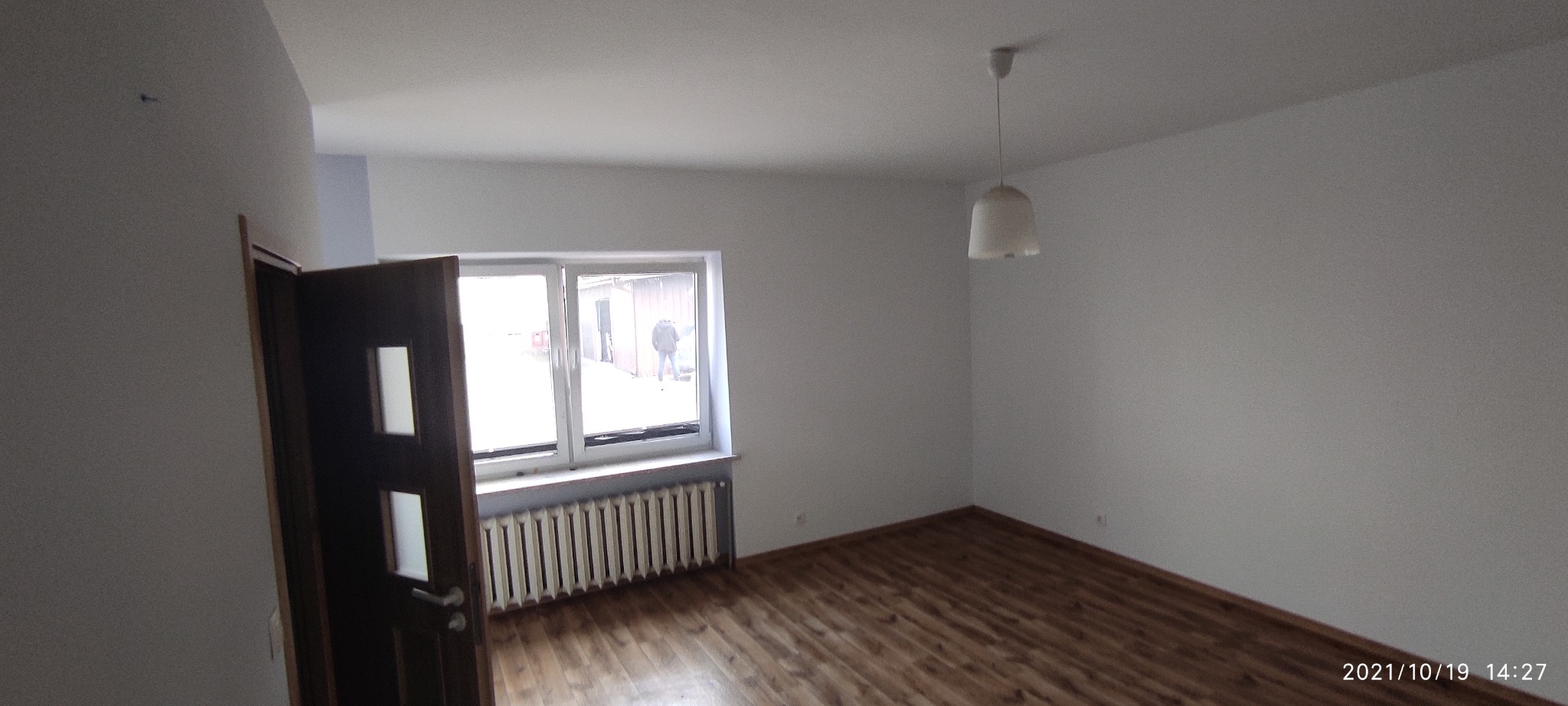 Pusty pokój z drewnianą podłogą, grzejnikiem pod oknem, otwartymi drzwiami z przeszkleniami i widokiem na zewnątrz. Świeżo pomalowane ściany w odcieniach bieli, surowy stan wykończenia.