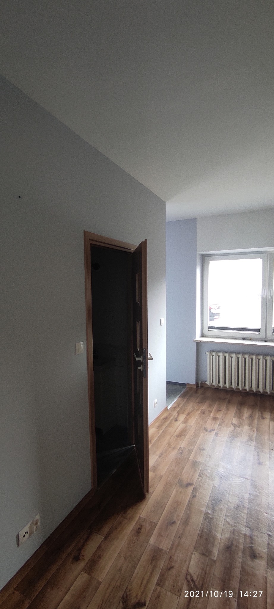 Wnętrze pokoju po malowaniu z drewnianą podłogą i częściowo otwartymi drewnianymi drzwiami, widok na grzejnik pod oknem.