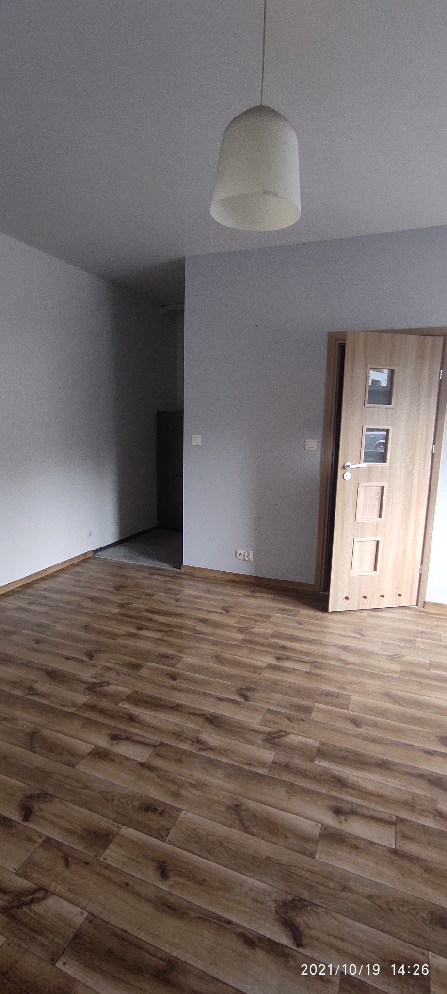 Pusty pokój z panelami podłogowymi w odcieniach brązu, szare ściany, drewniane drzwi z trzema okienkami i biała lampa wisząca