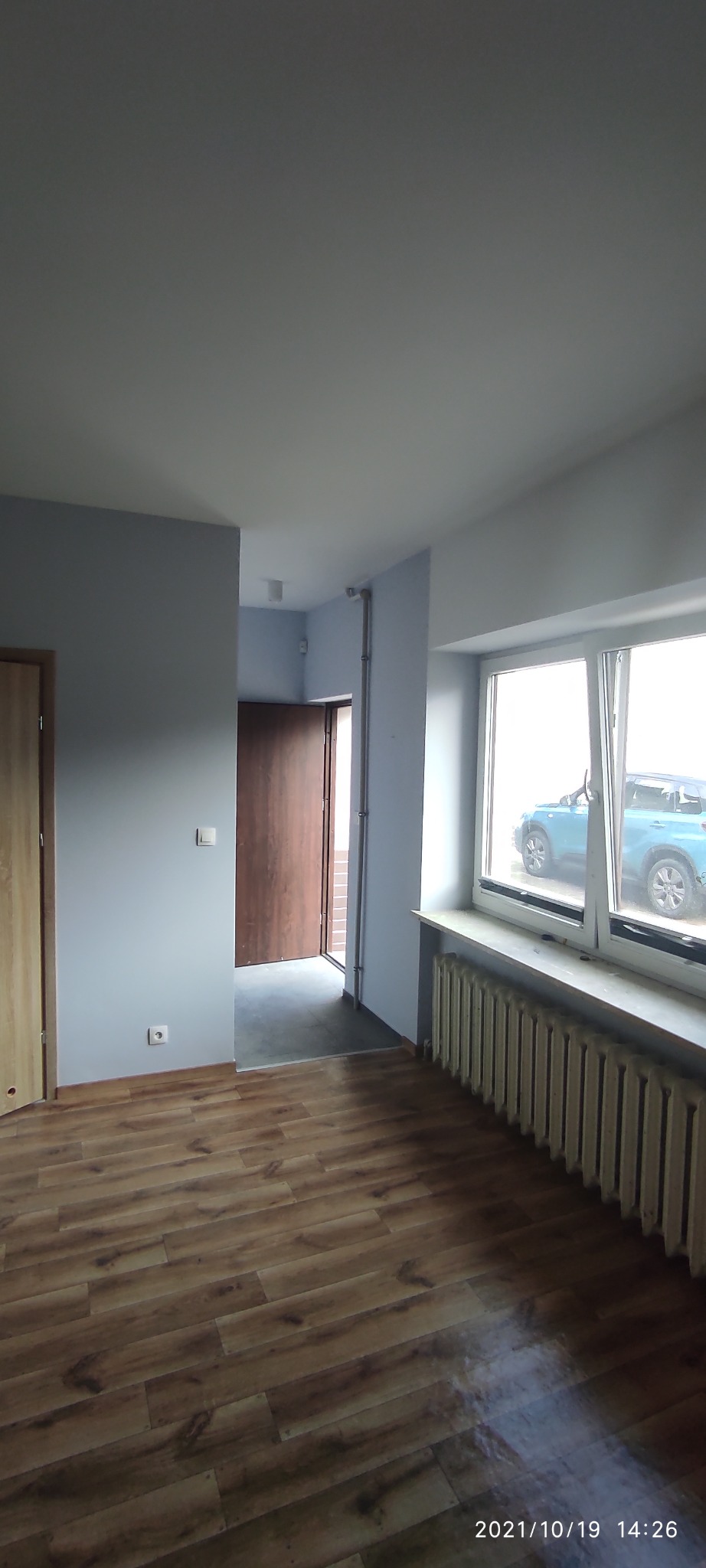 Wnętrze mieszkania po remoncie: świeżo pomalowane ściany na szaro, drewnopodobne panele podłogowe, grzejnik pod oknem, widok na korytarz z drewnianymi drzwiami wejściowymi.