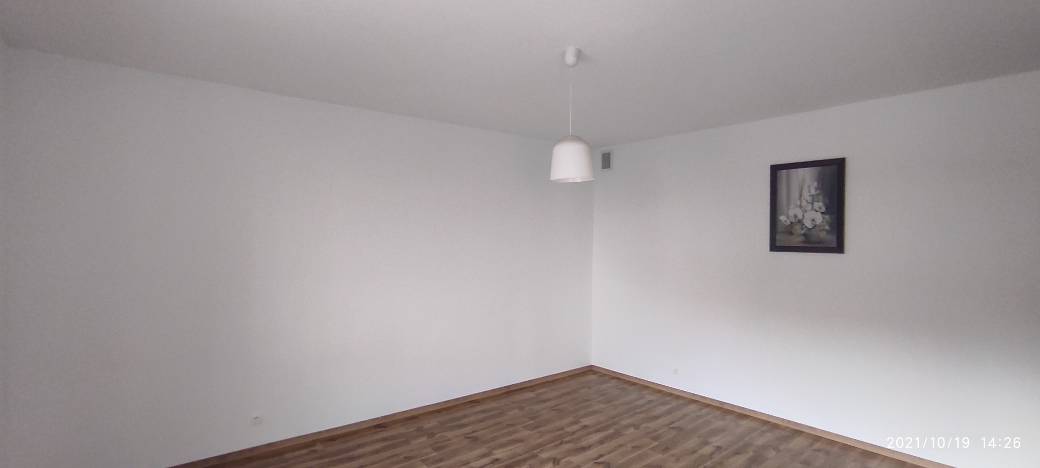 Pusty pokój z białymi ścianami, drewnianą podłogą, wiszącą lampą i obrazem z kwiatami w ciemnej ramie.