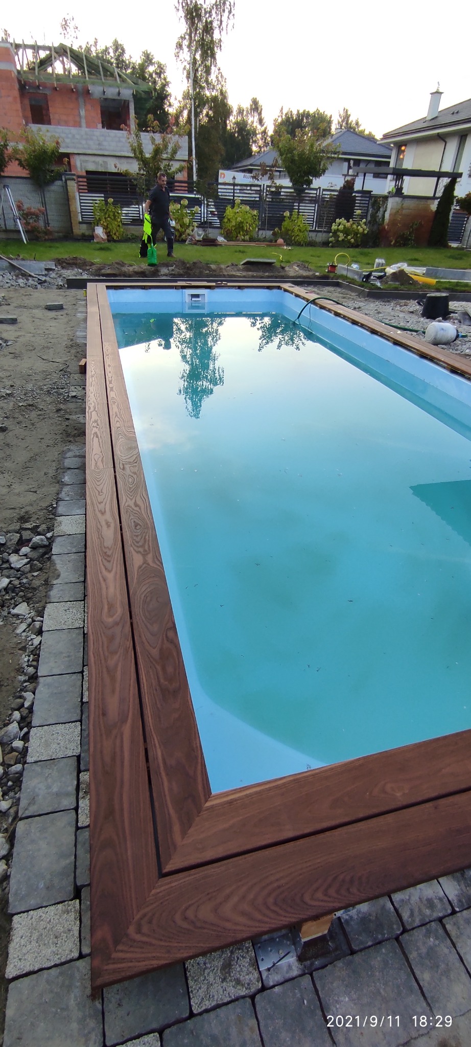 Niebieski basen z drewnianym obramowaniem w trakcie budowy, widoczny pracownik i dom w tle.