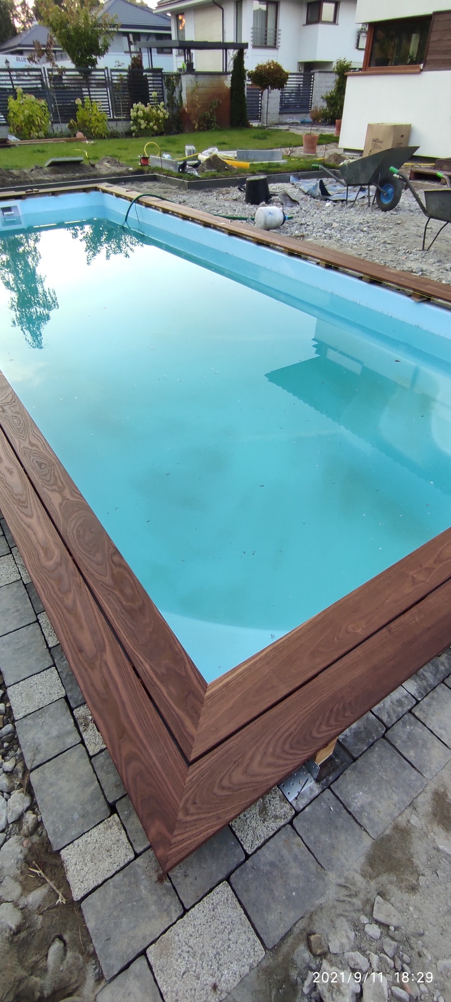 Prostokątny basen z drewnianym obramowaniem w trakcie budowy, widoczne narzędzia i materiały budowlane w tle.