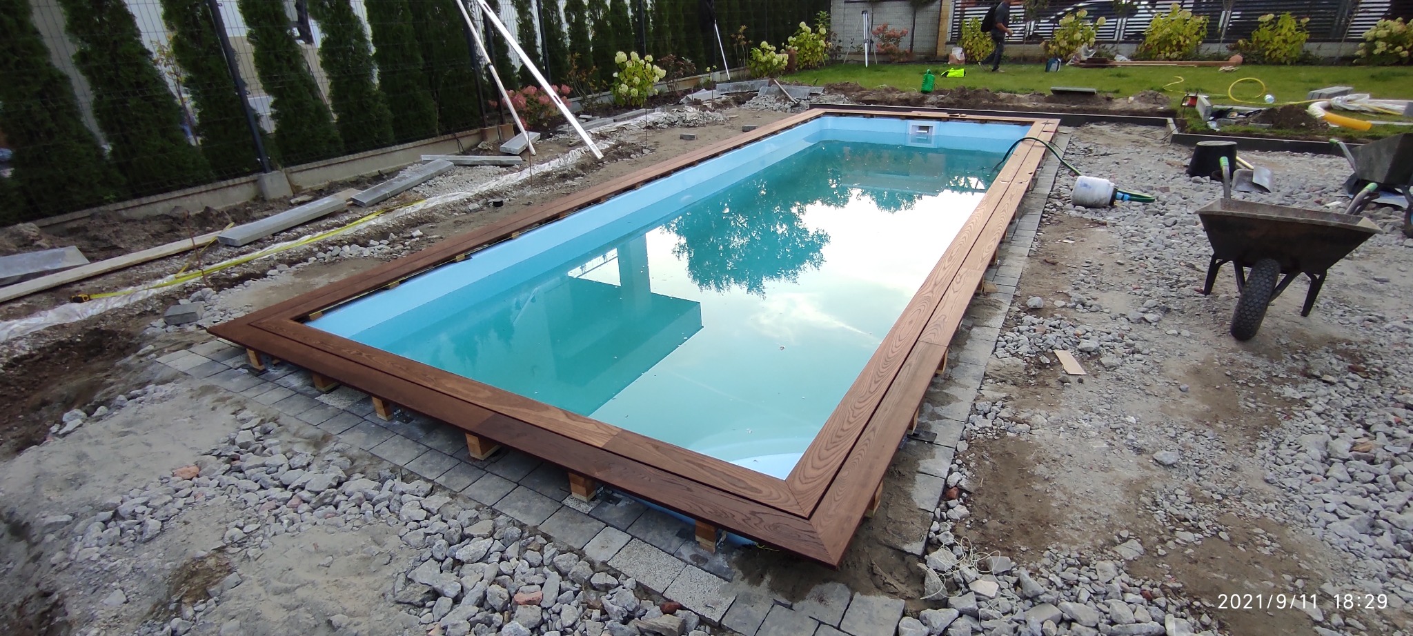 Prostokątny basen z jasną wodą i drewnianą ramą, w trakcie budowy, widoczne materiały budowlane i narzędzia w otoczeniu.