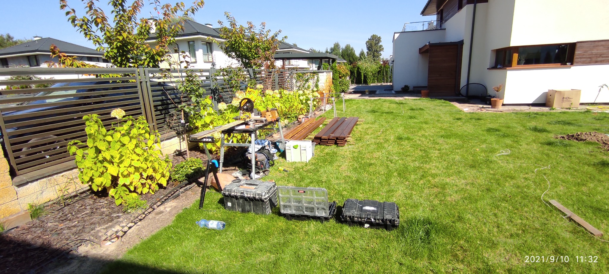 Przygotowania do montażu tarasu kompozytowego: deski, narzędzia i skrzynki na trawniku przed domem z nowoczesną architekturą.