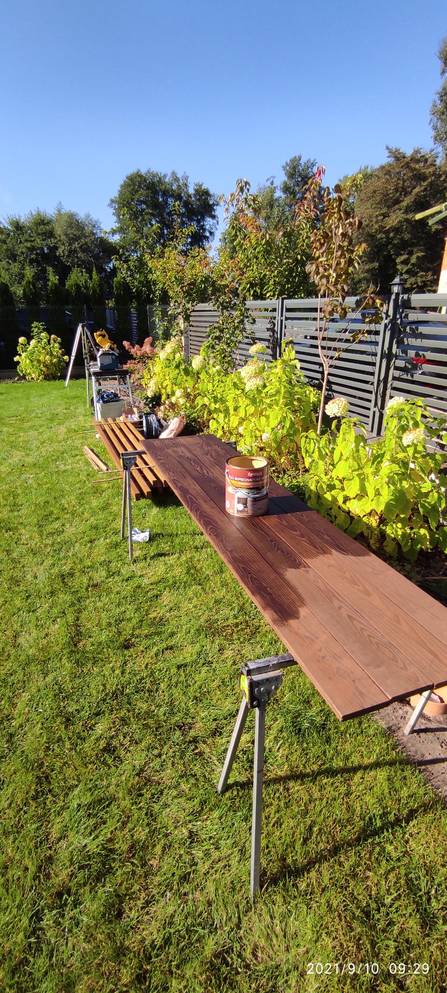 Impregnowana deska tarasowa na stojakach w ogrodzie, częściowo pokryta olejem, puszka z olejem impregnacyjnym widoczna na desce, w tle zielona trawa i krzewy.