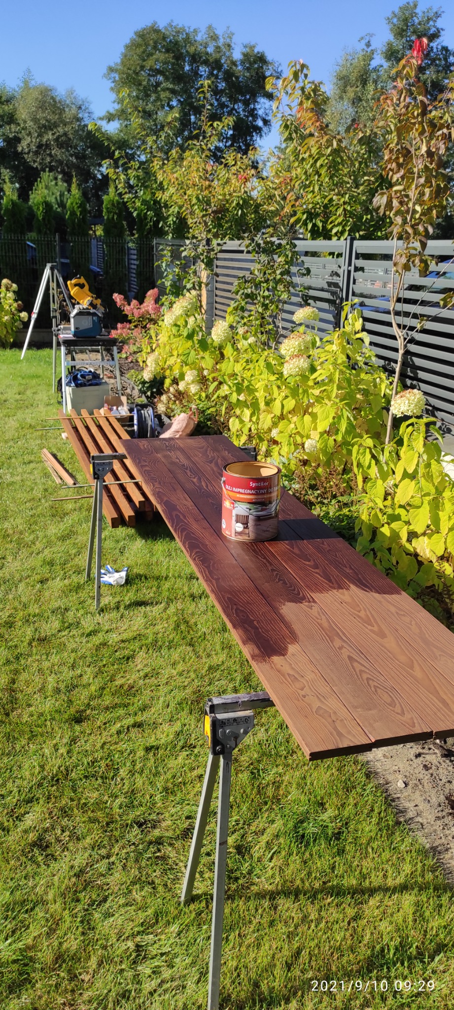 Impregnacja desek tarasowych na zewnątrz, widoczna puszka z olejem impregnacyjnym, część desek już zaimpregnowana, na trawie, w tle ogród.