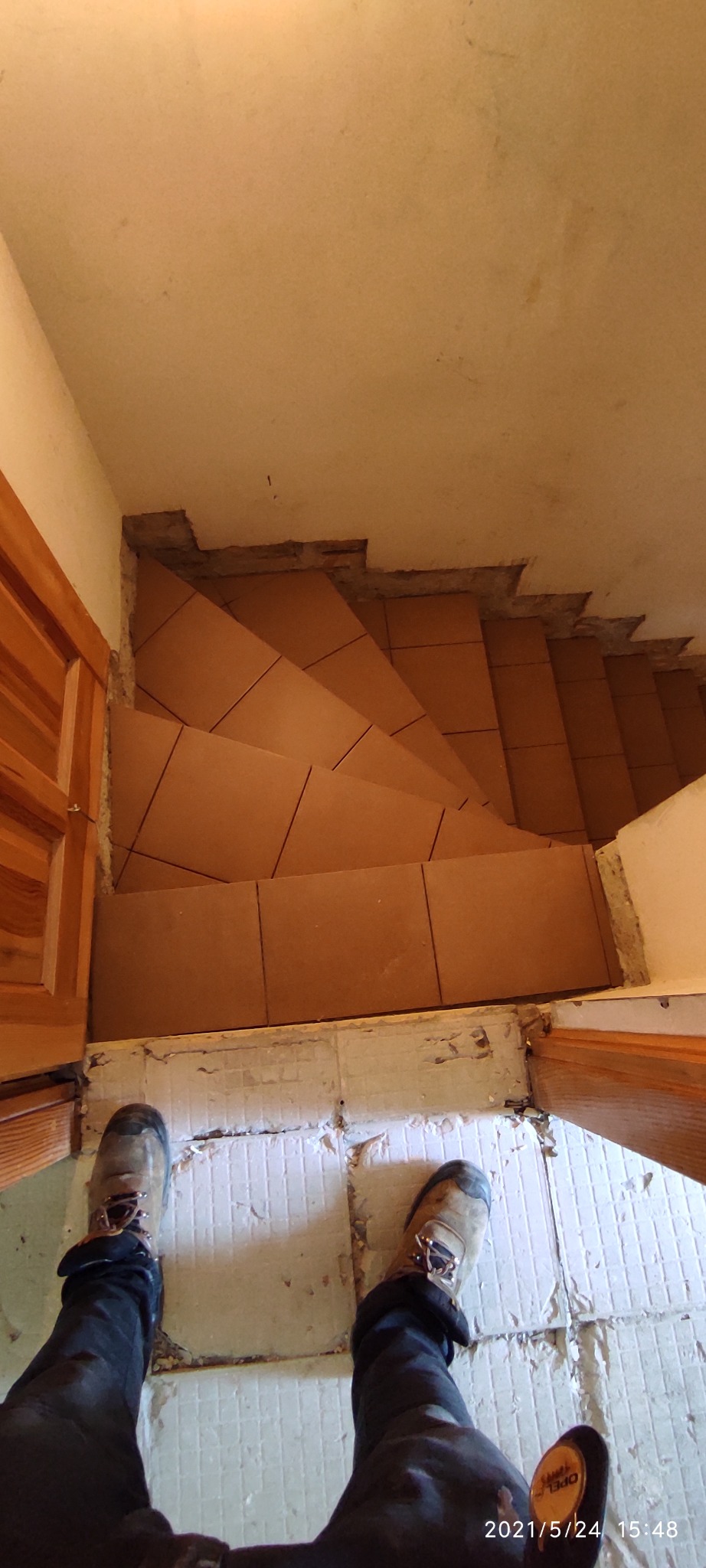 Montaż brązowych płytek na schodach w trakcie prac wykończeniowych, widok z góry na schody i robotnika w butach roboczych.