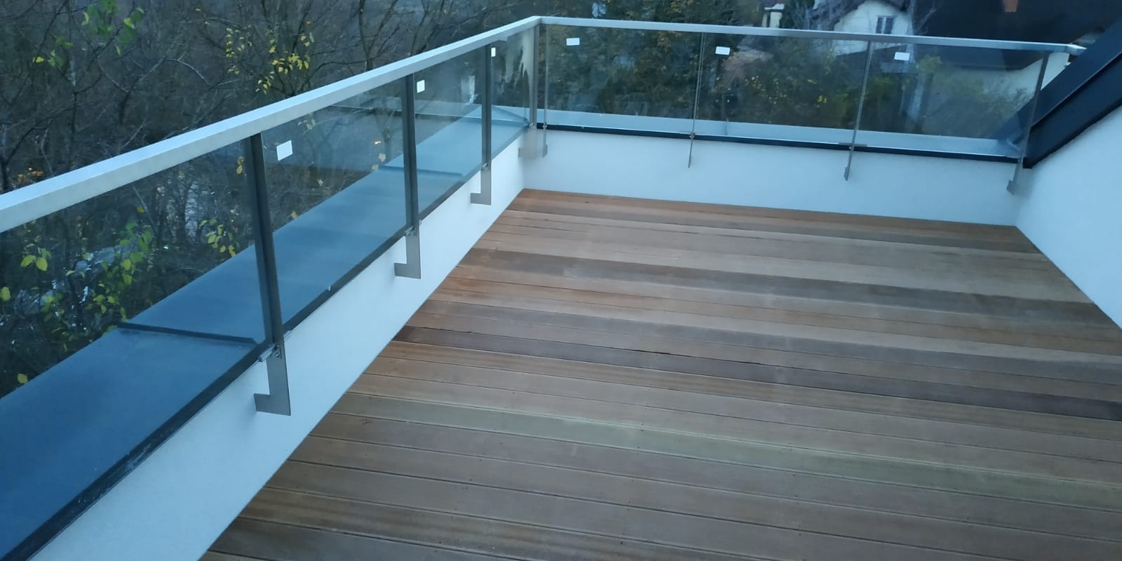 Drewniany taras z balustradą ze szkła i metalu, widok z góry, jesienne drzewa w tle.