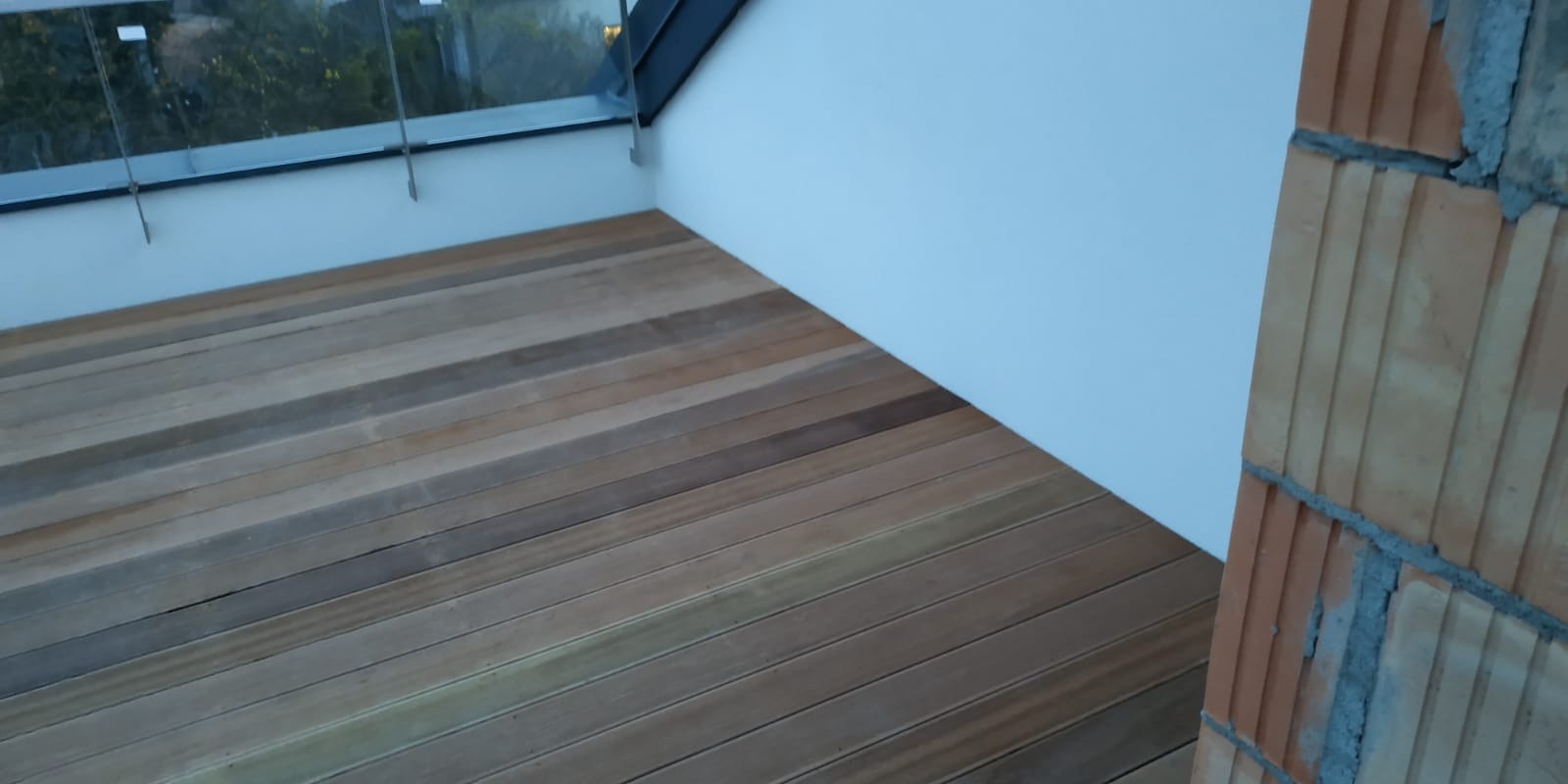 Nowoczesny taras z drewnianą podłogą, szklaną balustradą i fragmentem murowanej ściany w surowym stanie.