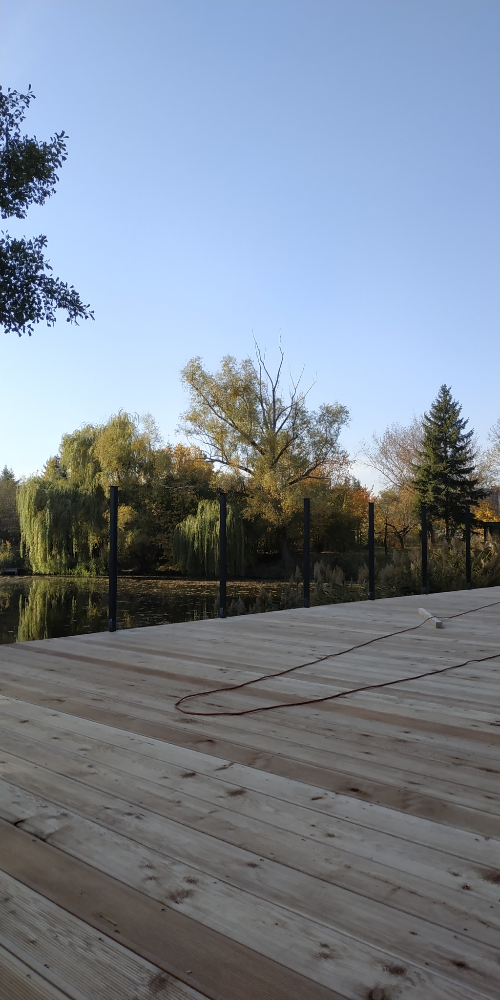 Nowy drewniany taras z czarnymi słupkami, widok na spokojne jezioro i jesienne drzewa w tle, kabel narzędziowy leżący na deskach.