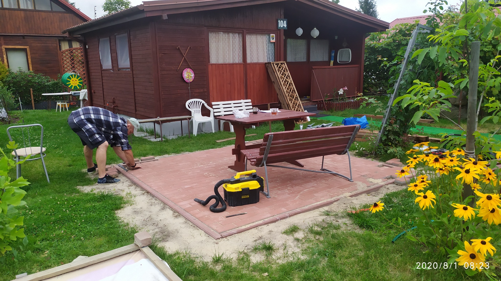 Układanie czerwonej kostki brukowej na tarasie przed drewnianym domkiem letniskowym, widoczny pracownik w kraciastej koszuli, stół z ławkami i odkurzacz przemysłowy.