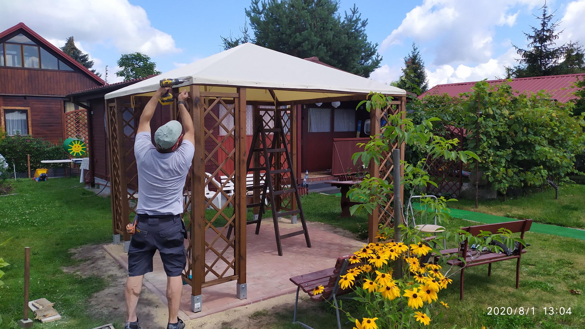 Monterażysta przykręca wkrętarką element drewnianej pergoli z białym dachem na tle ogrodu z kwitnącymi rudbekiami i domkiem letniskowym.