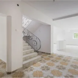 Jasne, przestronne wnętrze domu z kafelkową podłogą w geometryczne wzory, schodami z kutą balustradą i widokiem na ogr&oacute;d przez okno w głębi.