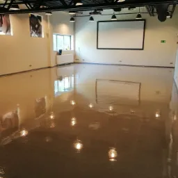 Wnętrze biura po gruntownym sprzątaniu, widoczna lśniąca, wypolerowana podłoga z refleksami świetlnymi, białe ściany z elementami dekoracyjnymi i oknem, sufit z widoczną instalacją oświetleniową...