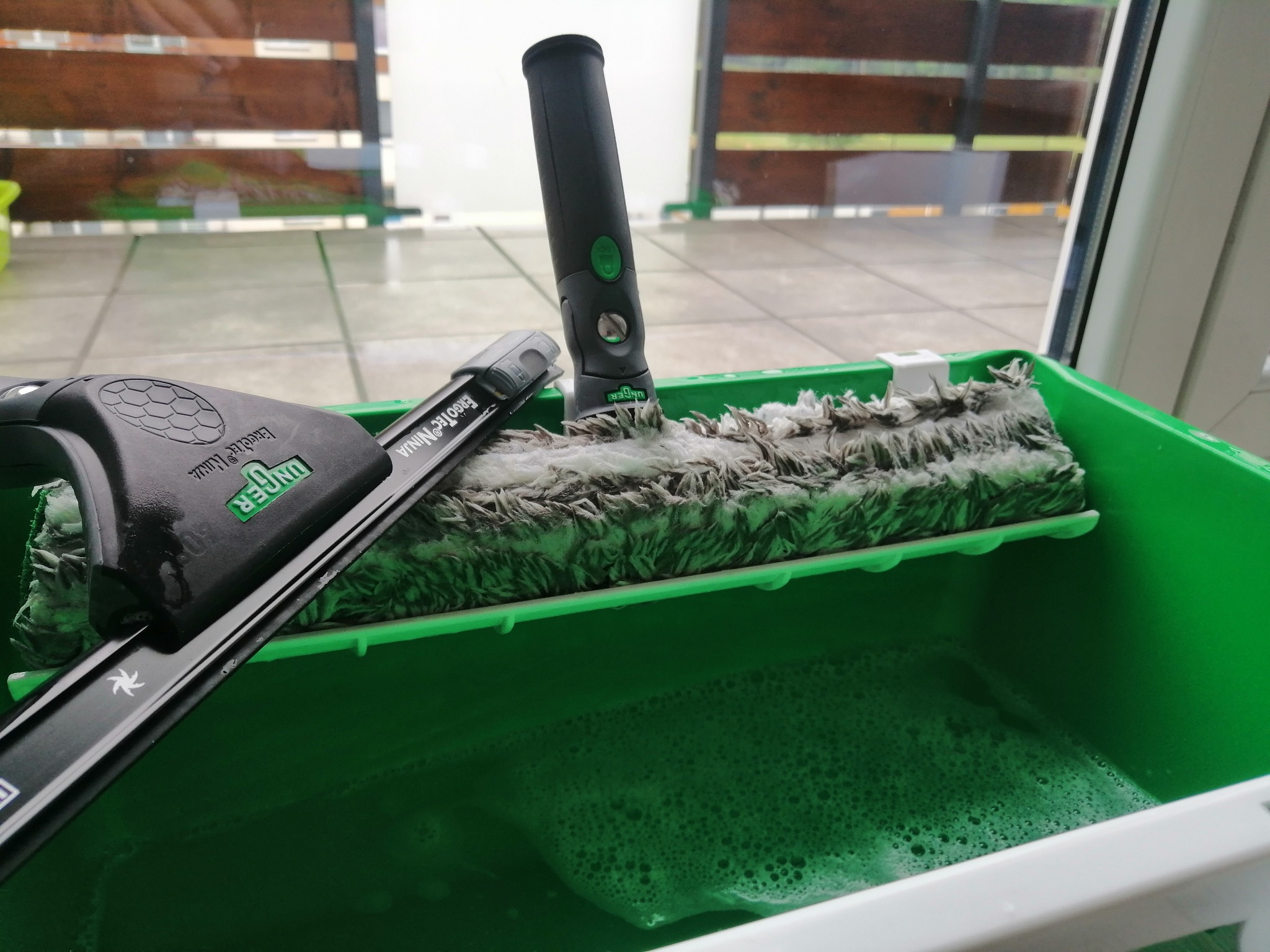 Zielone wiadro z wodą z mydłem i narzędziami do mycia okien marki Unger: mop z mikrofibry ErgoTec Ninja i ściągaczka ErgoTec Ninja, widoczne na tle okna z drewnianymi żaluzjami.