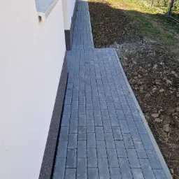 Świeżo ułożony chodnik z szarej kostki brukowej przylegający do białej ściany budynku, z widoczną ziemią przygotowaną pod dalsze prace ogrodowe.