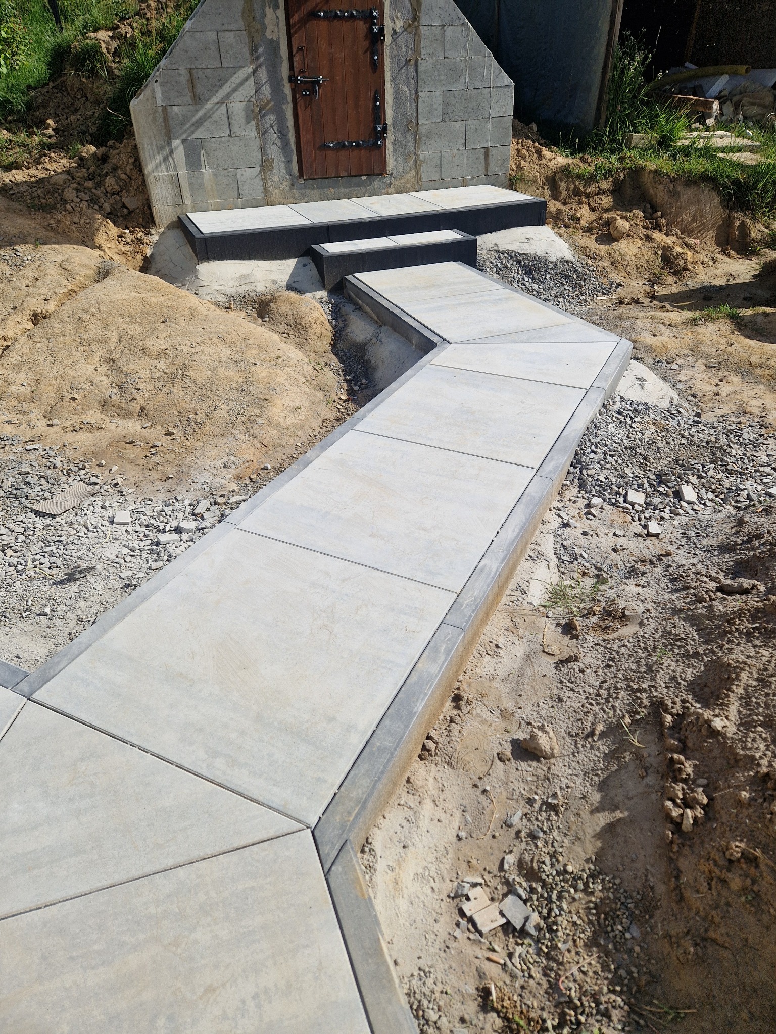 Ścieżka z jasnoszarej kostki brukowej prowadząca do wejścia piwnicy z drewnianymi drzwiami i betonowymi schodami, otoczona ziemią i gruzem.