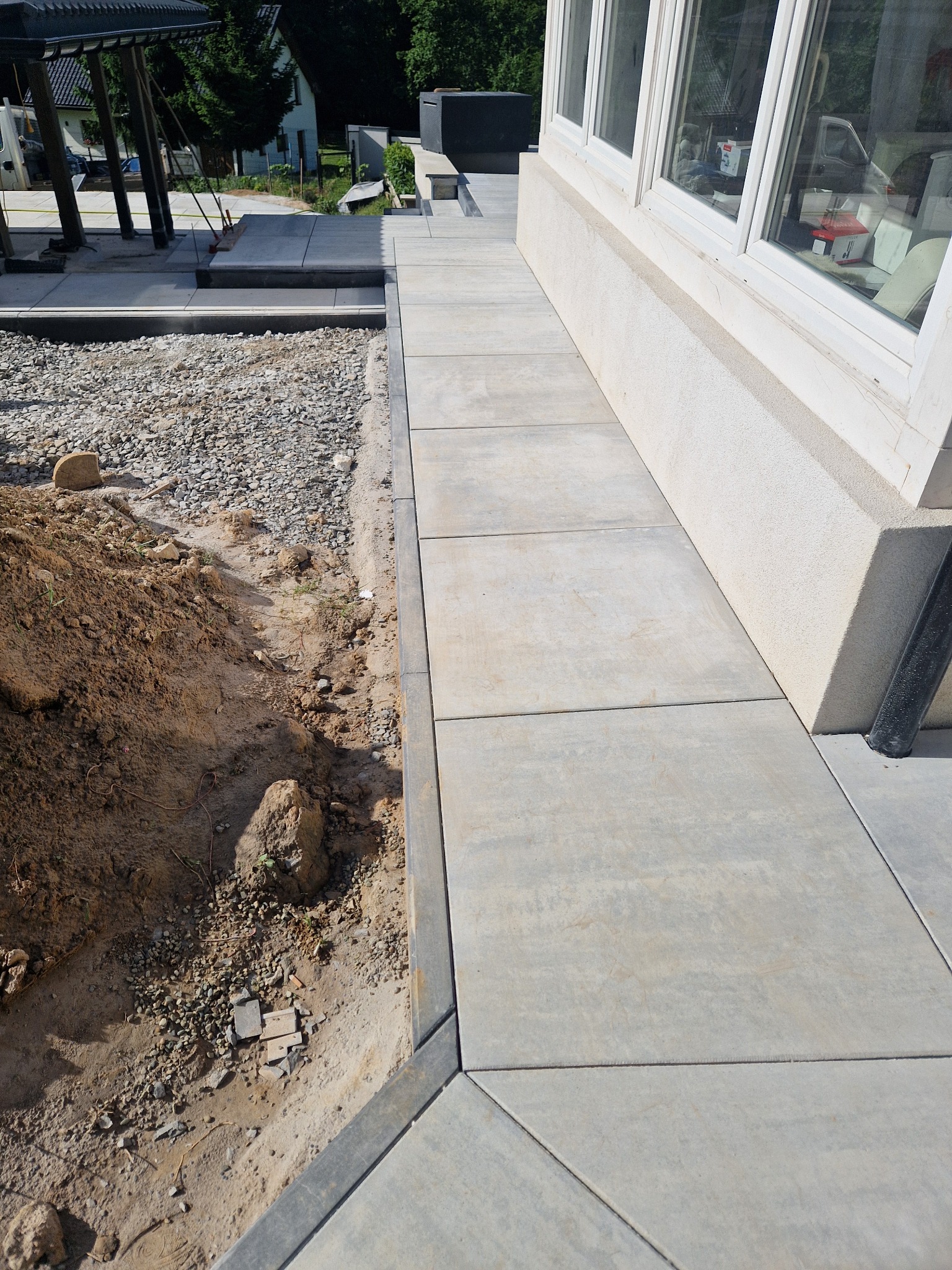 Świeżo ułożony chodnik z szarych płyt betonowych przylegający do ściany budynku, z widocznym fragmentem wykopu wypełnionego żwirem i ziemią po jednej stronie.