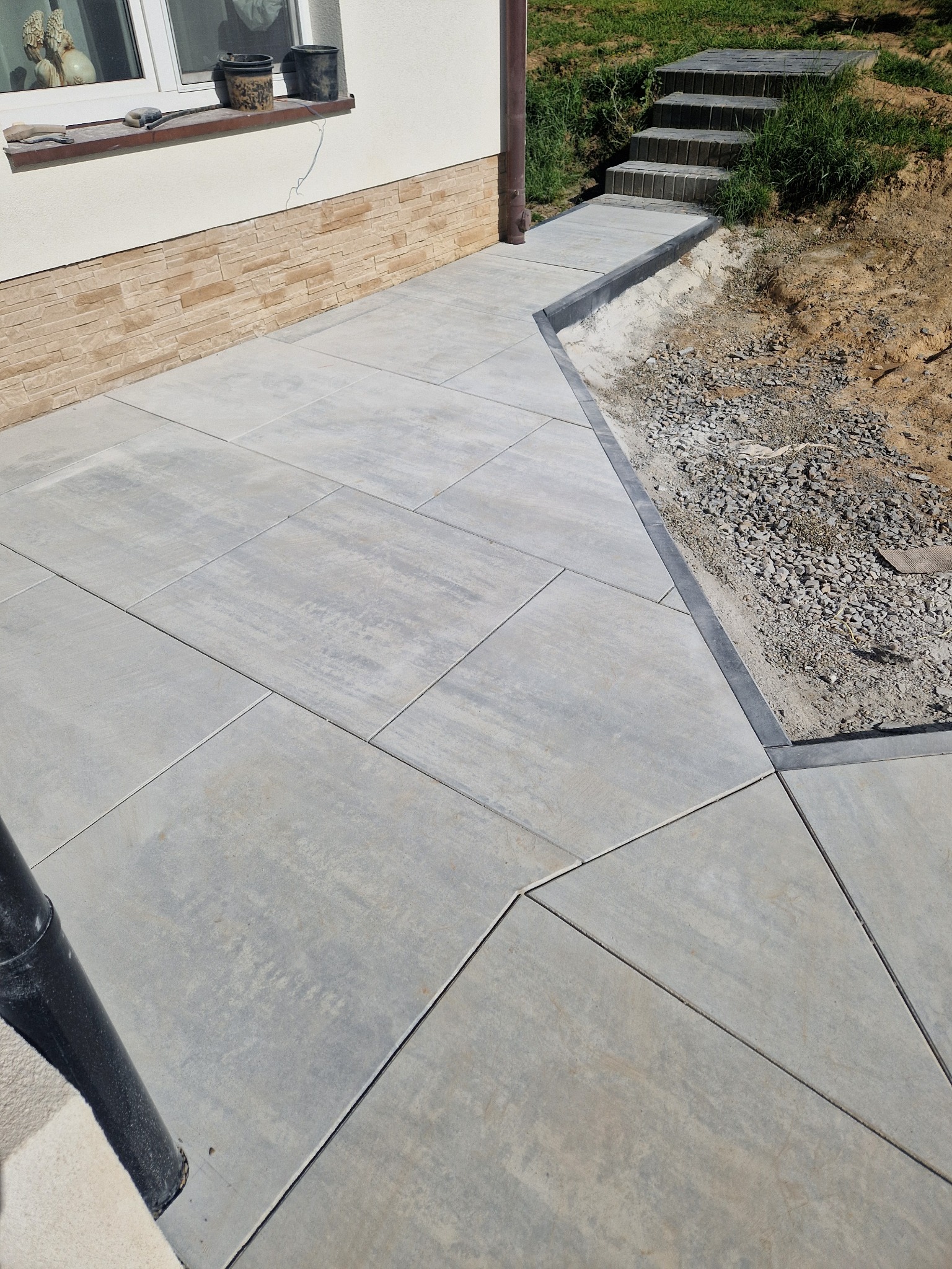 Nowo ułożona, szara nawierzchnia z dużych płyt betonowych prowadząca do schodów z kostki, widoczna rynna spustowa przy ścianie budynku.