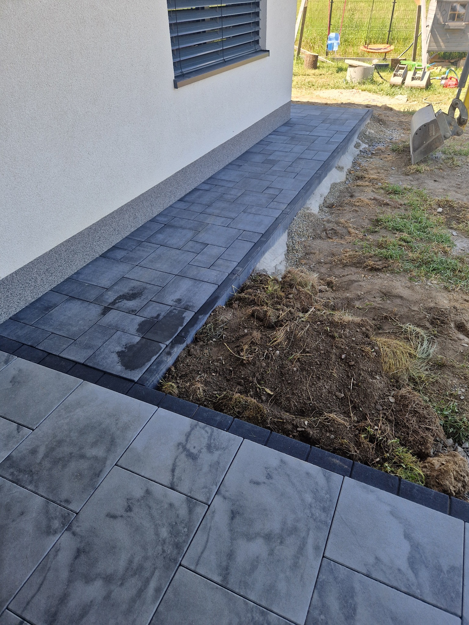 Świeżo ułożona, ciemnoszara kostka brukowa przy ścianie domu z widocznym fragmentem wykopu i łyżką koparki w tle.