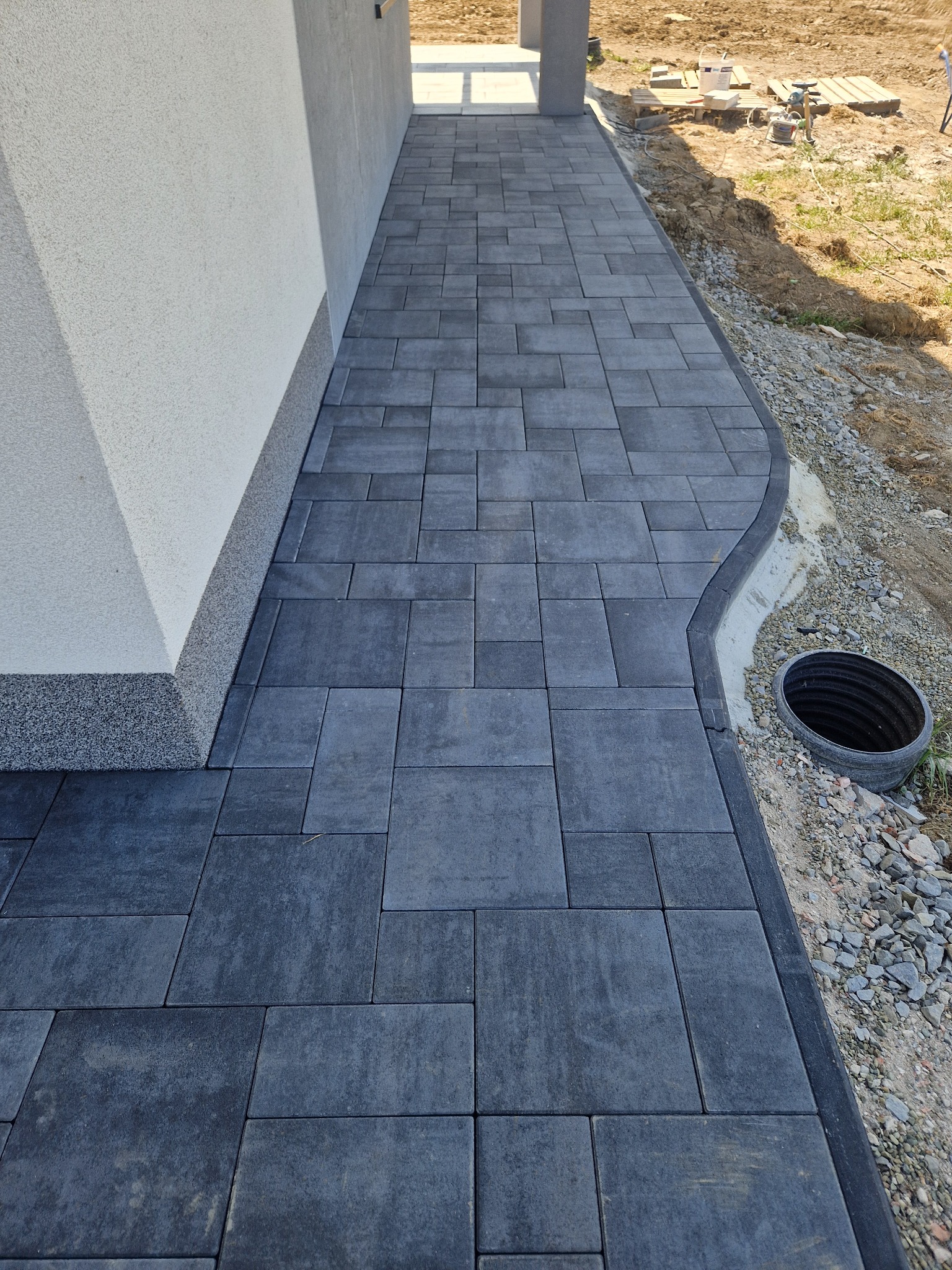 Ścieżka z ciemnoszarej kostki brukowej ułożona przy białym budynku, obrzeże z ciemniejszej kostki, widoczny wykop i rura kanalizacyjna.
