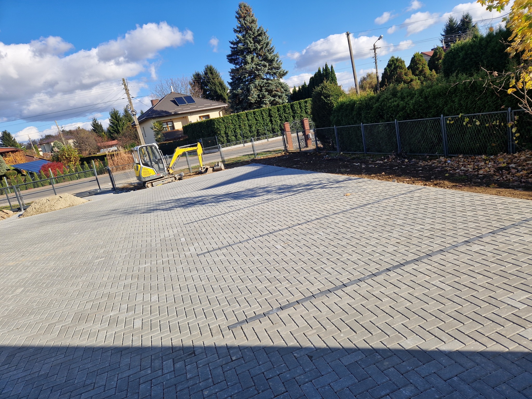 Ułożona szara kostka brukowa na posesji, mała koparka na skraju placu, ogrodzenie siatkowe, dom z panelami słonecznymi w tle pod błękitnym niebem z chmurami.
