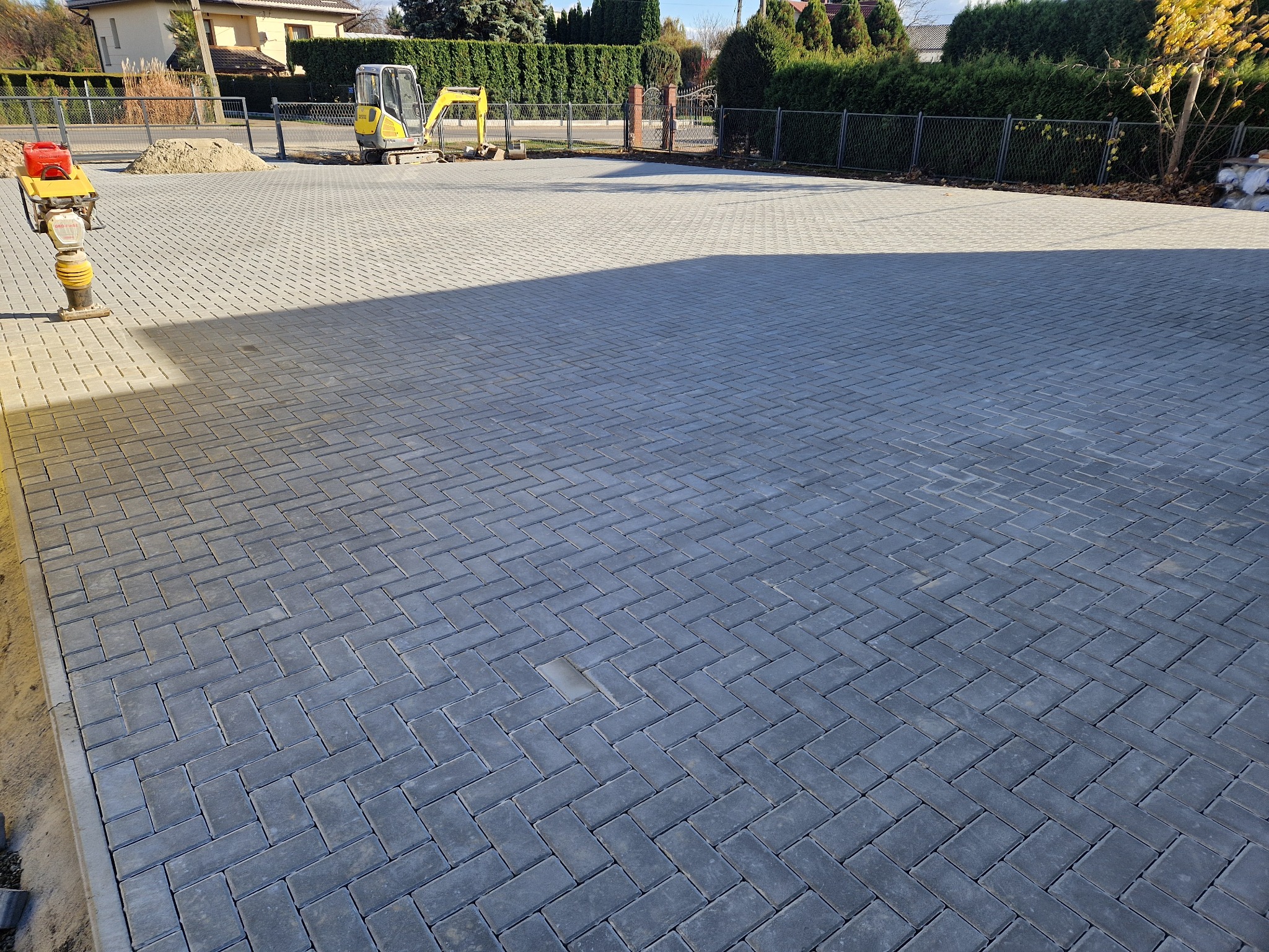 Ukończona powierzchnia z szarej kostki ułożonej w jodełkę, z lekkim cieniem i sprzętem budowlanym w tle, widok z góry.