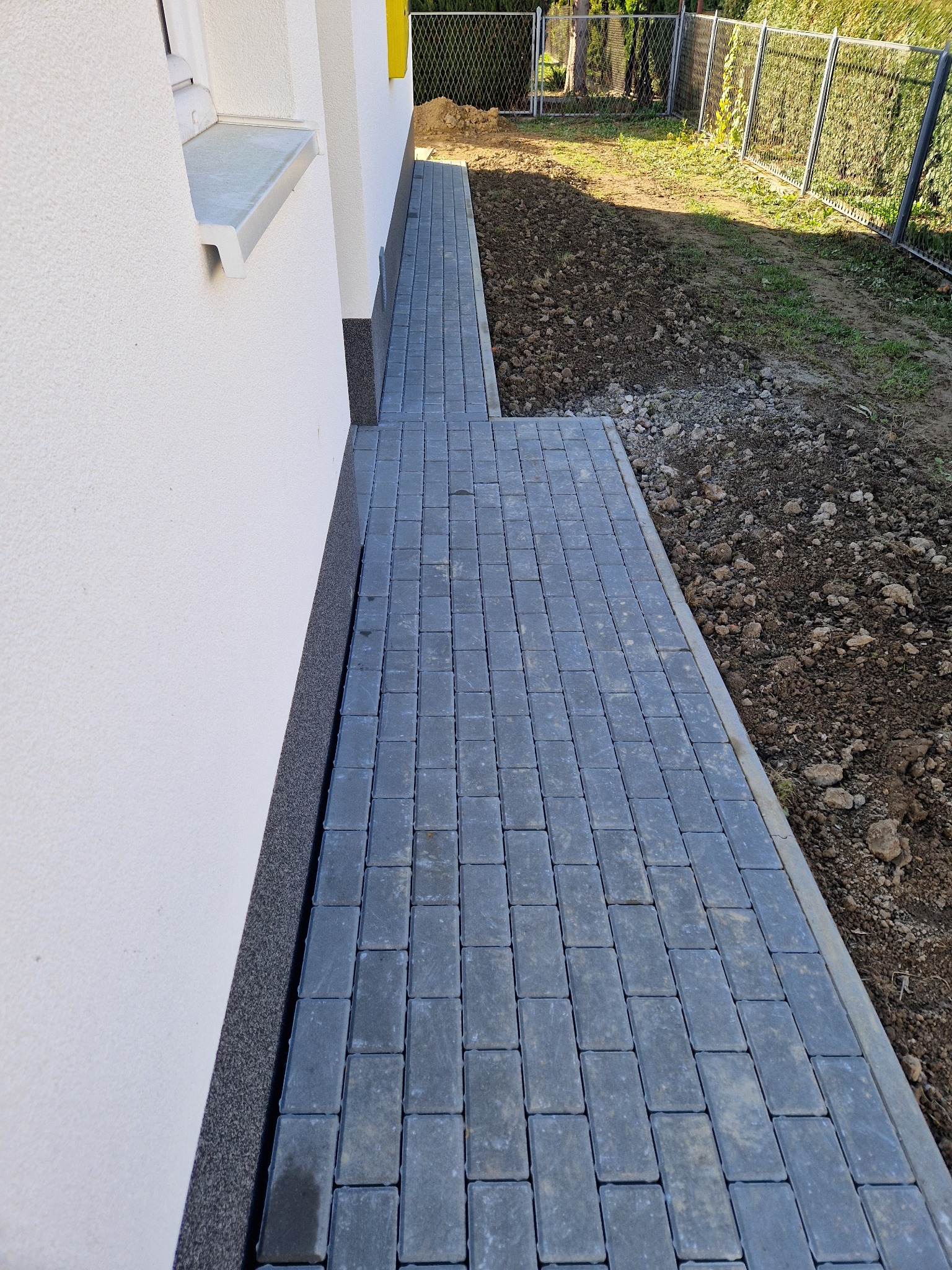 Świeżo ułożony chodnik z szarej kostki brukowej przylegający do białej ściany budynku, z widoczną ziemią przygotowaną pod dalsze prace ogrodowe.