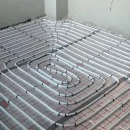 Białe rury ogrzewania podłogowego, równomiernie rozłożone i przymocowane do siatki na podłodze w surowym wnętrzu, widok z góry.
