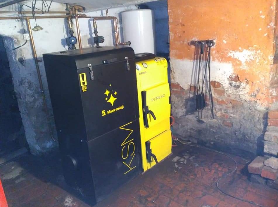 Czarno-żółty piec KSM Pereko na paliwo stałe, klasa emisji 5, zainstalowany w piwnicy z widocznymi rurami instalacji grzewczej i elementami kotłowni.