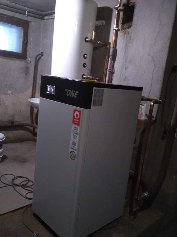 Nowoczesny piec grzewczy 'ONE' z podłączonymi miedzianymi rurami w piwnicy z betonową ścianą i małym oknem.