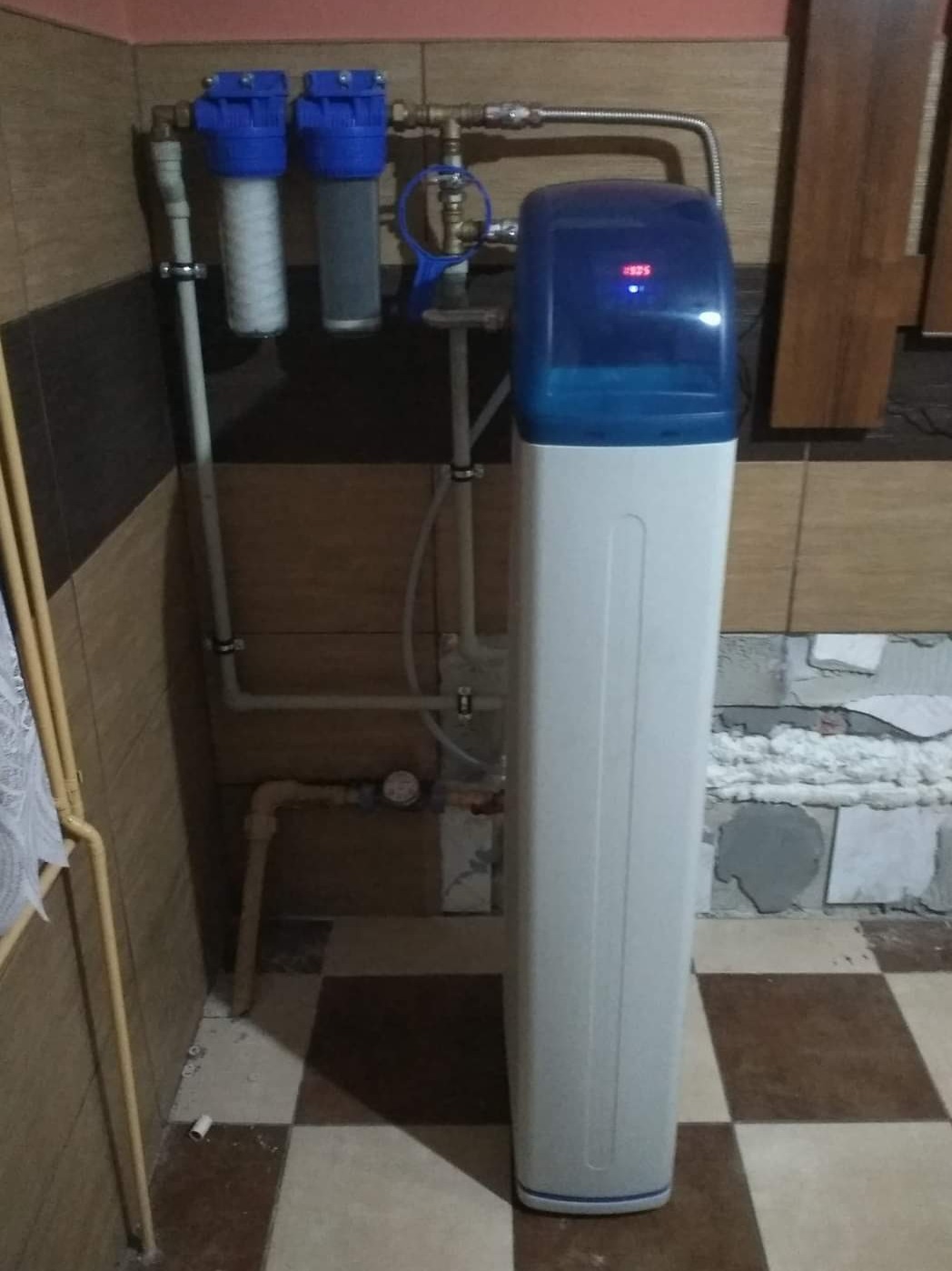 Filtr do wody z niebieską górną częścią i białym korpusem, zainstalowany w pomieszczeniu z brązowymi i beżowymi płytkami, widoczne rury i dodatkowe filtry wstępne na ścianie.