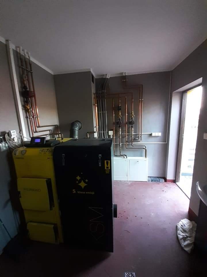 Kotłownia z dwoma kotłami: żółtym i czarnym, z widocznymi rurami miedzianymi i białym szafką w tle, podłoga w kolorze bordo.